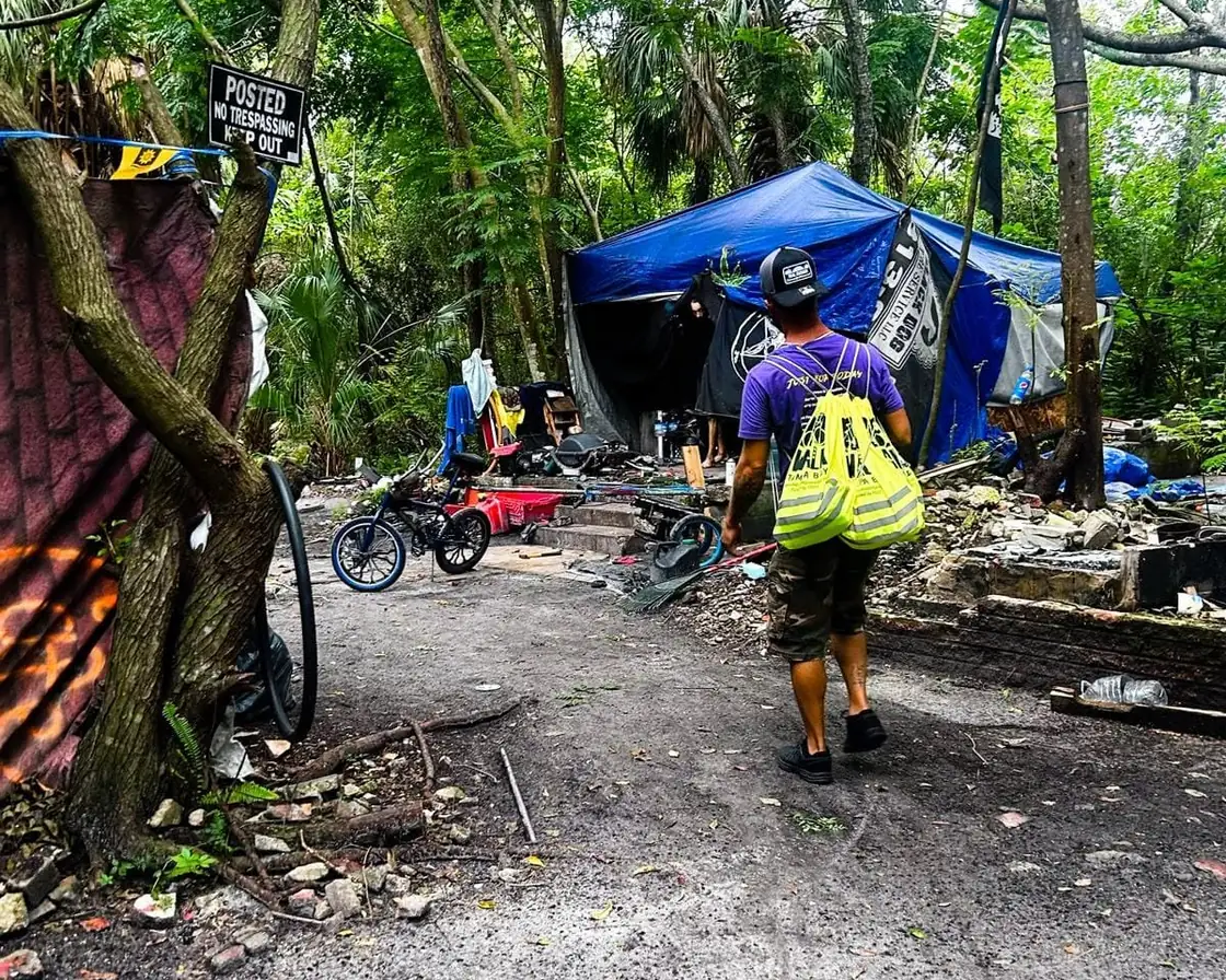 A man in a purple shirt carries yellow drawstring bags into a wooded encampment, toward a shelter of blue tarps